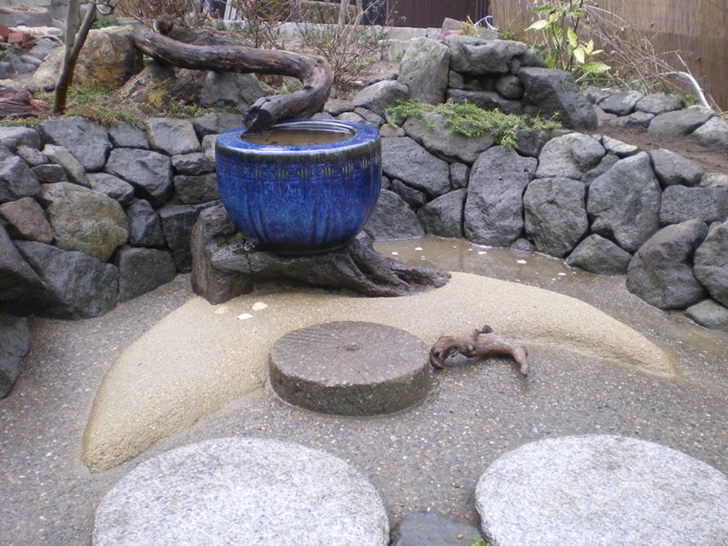 既存の火鉢を水鉢として背景に石を積み、水鉢の下に三日月形の洗い出しを設けた庭